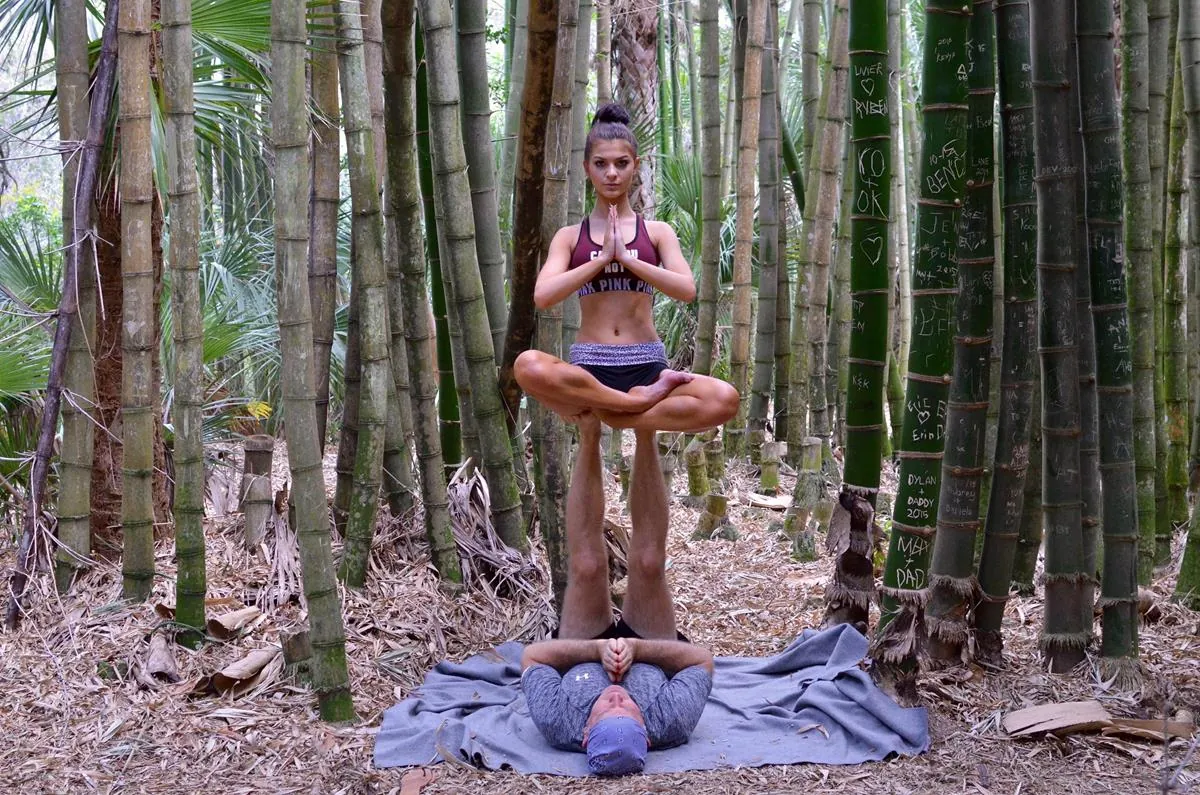 An acro yoga flyer balances in lotus pose on her base's feet in a bamboo grove