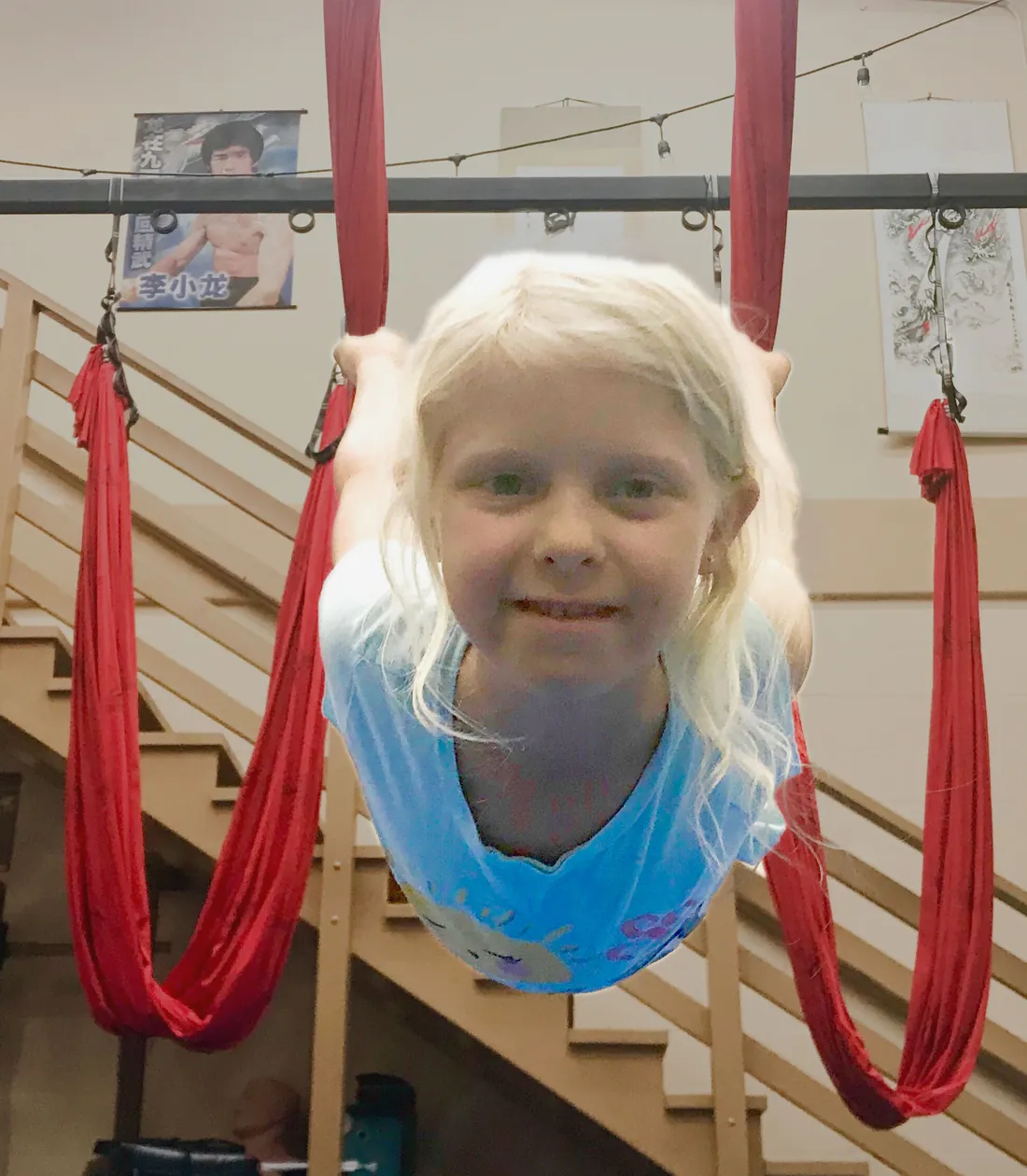 A smiling child hangs upside down in an aerial hammock